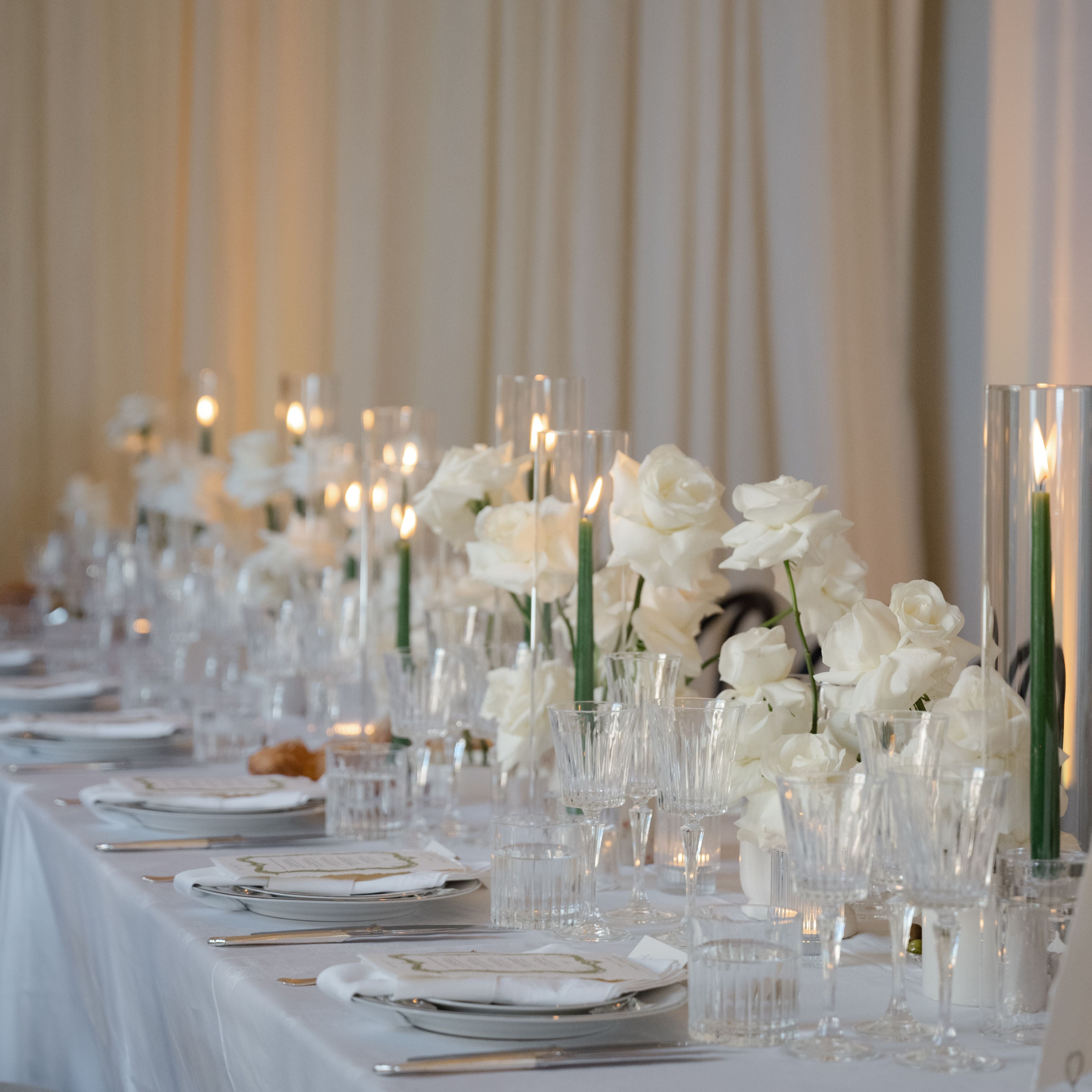 Elegant table setting with white flowers, candles, and silverware against a beige curtain background.
